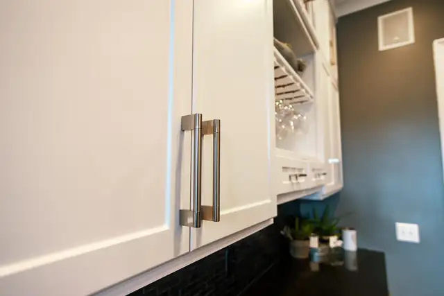 Close-up of white satin Shaker cabinets with long bar pulls beside a wine glass rack and dark tile backsplash